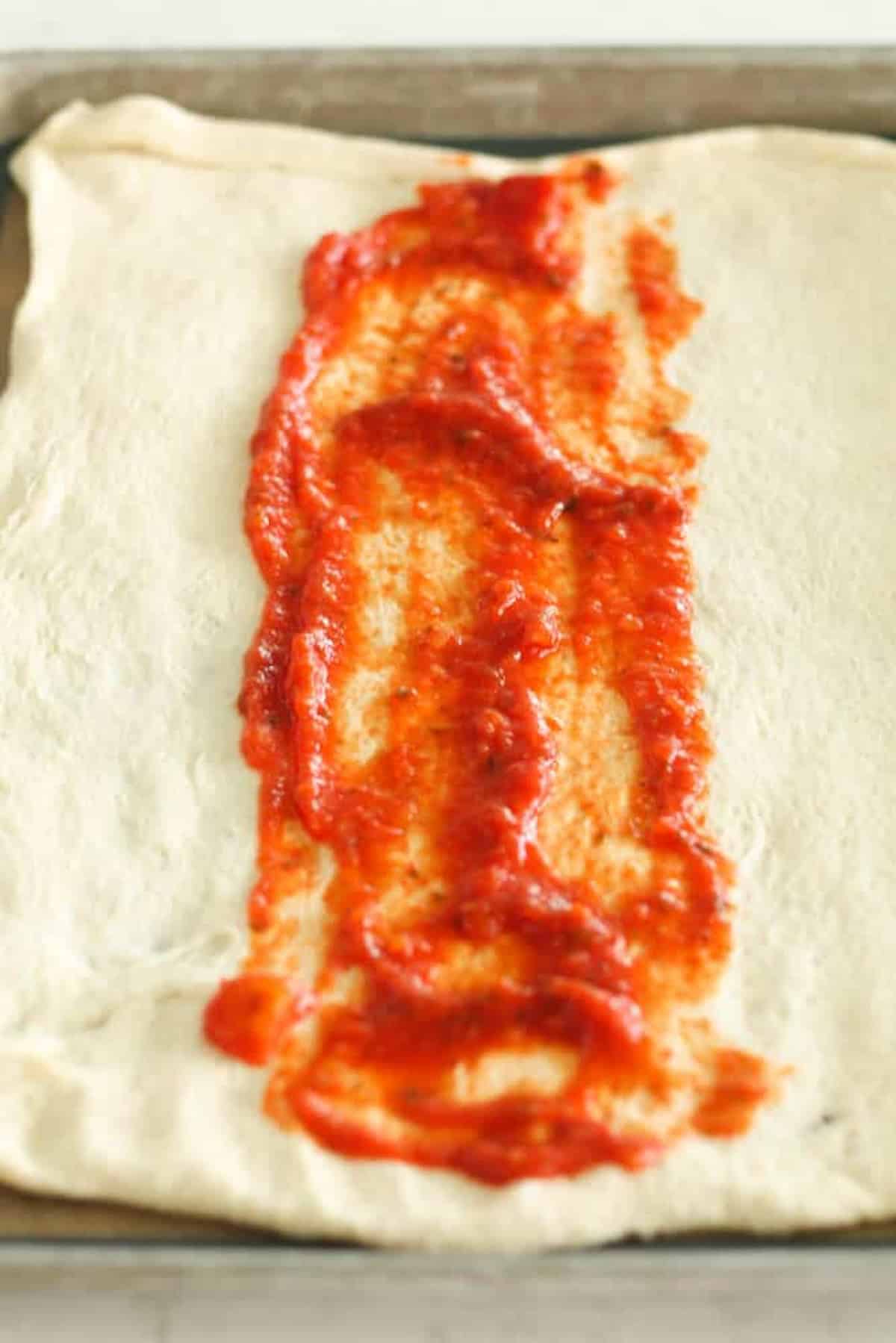 Rectangular pizza dough with a strip of tomato sauce spread down the center on a baking tray.