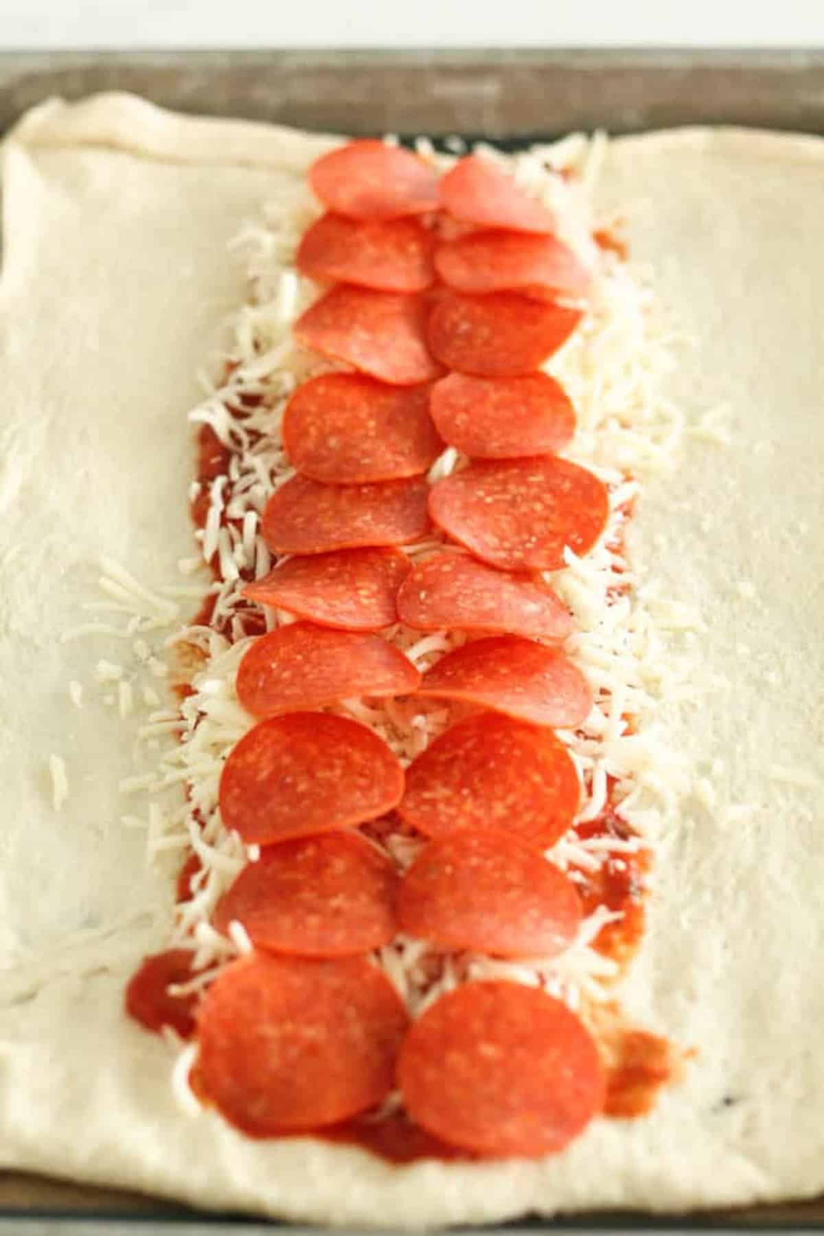 Pizza dough topped with sauce, shredded cheese, and a row of pepperoni slices down the middle, ready to be folded or rolled.