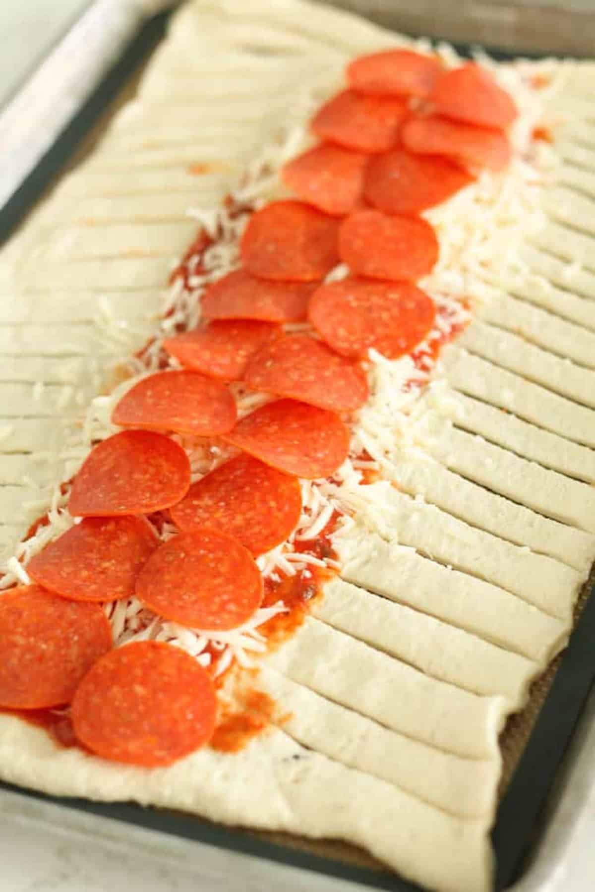 Unbaked braided pizza dough topped with sliced pepperoni, tomato sauce, and shredded cheese, laid out on a baking sheet.