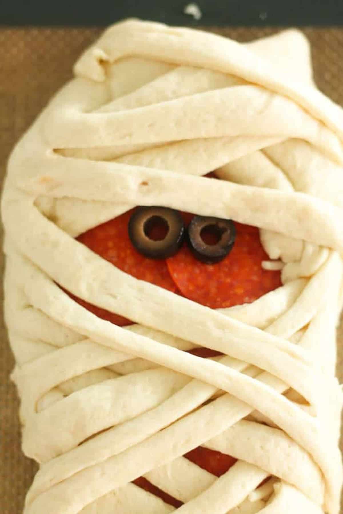 A bread dough made to resemble a wrapped mummy, with slices of olives peeking through for eyes and pepperoni visible underneath.