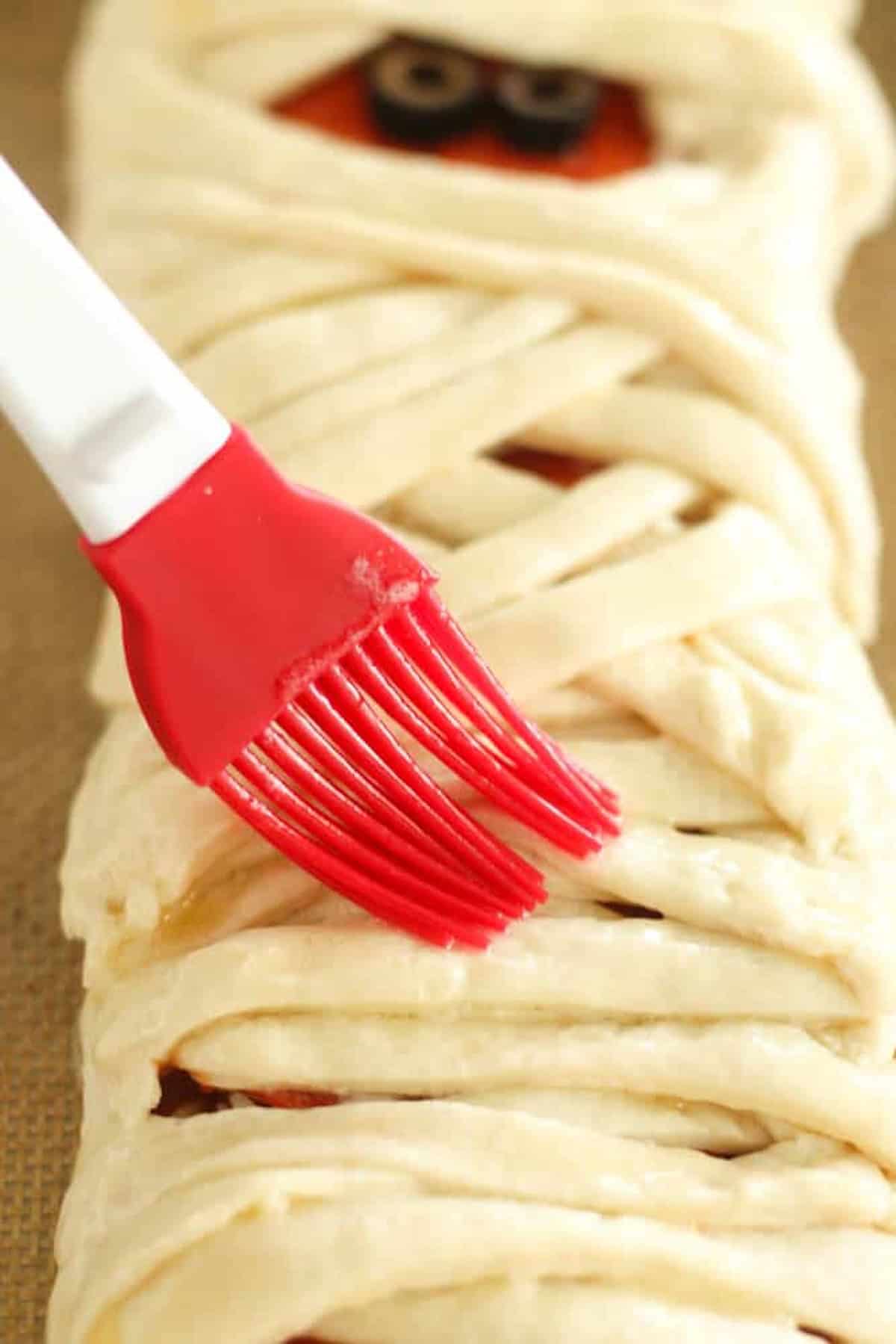 A red silicone brush is applying an egg wash to a pastry designed to look like a mummy, with strips of dough crisscrossed over a baked surface with small black eyes visible.