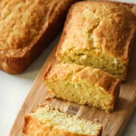 Two loaves of golden brown bread are placed on a wooden cutting board, with one loaf partially sliced into pieces, revealing a moist, crumbly texture inside.