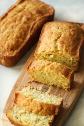 Two loaves of golden brown bread are placed on a wooden cutting board, with one loaf partially sliced into pieces, revealing a moist, crumbly texture inside.