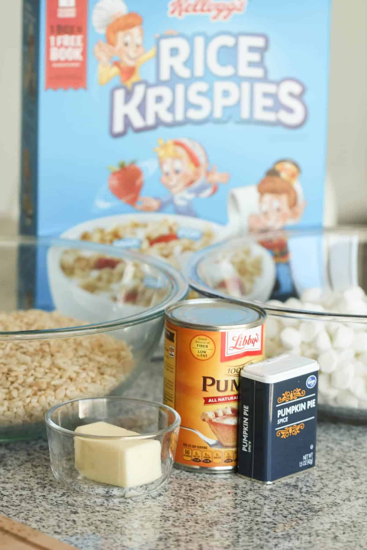 Ingredients for making Rice Krispies treats including a box of Rice Krispies, a glass bowl of Rice Krispies, canned pumpkin, pumpkin pie spice, butter, and marshmallows.