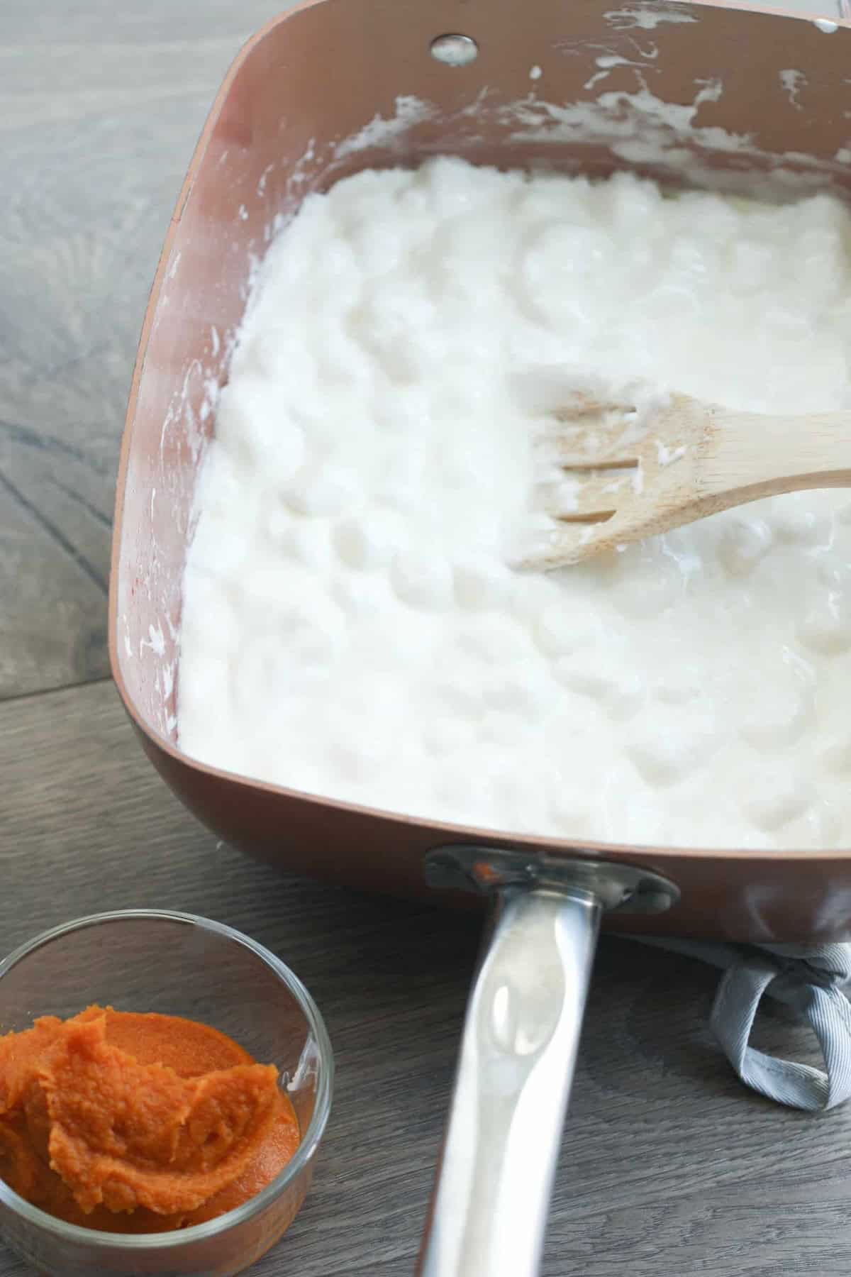 A square pan filled with a white, fluffy mixture being stirred with a wooden spoon. Next to the pan, there's a small glass bowl containing an orange paste.