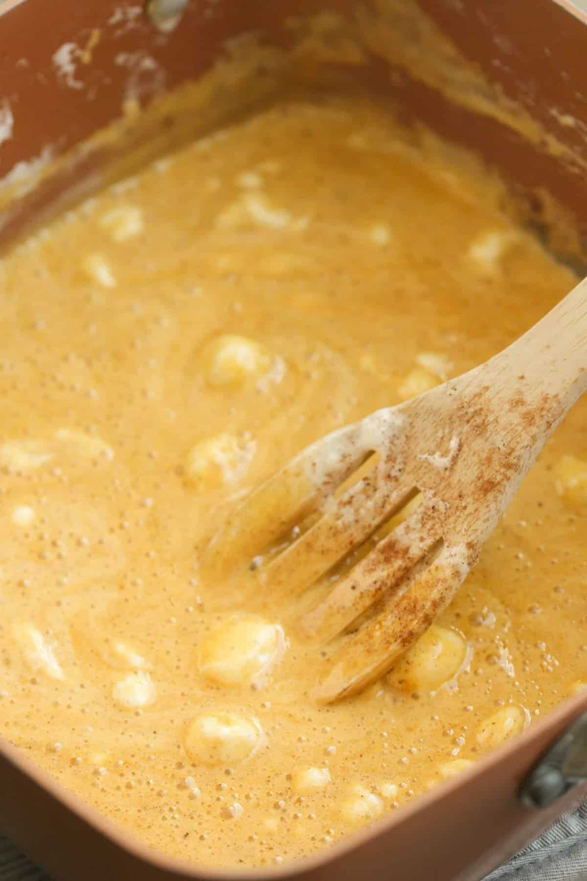 A glass bowl containing a mixture of melted marshmallow, cereal, and possibly pumpkin puree being stirred with a wooden spoon.