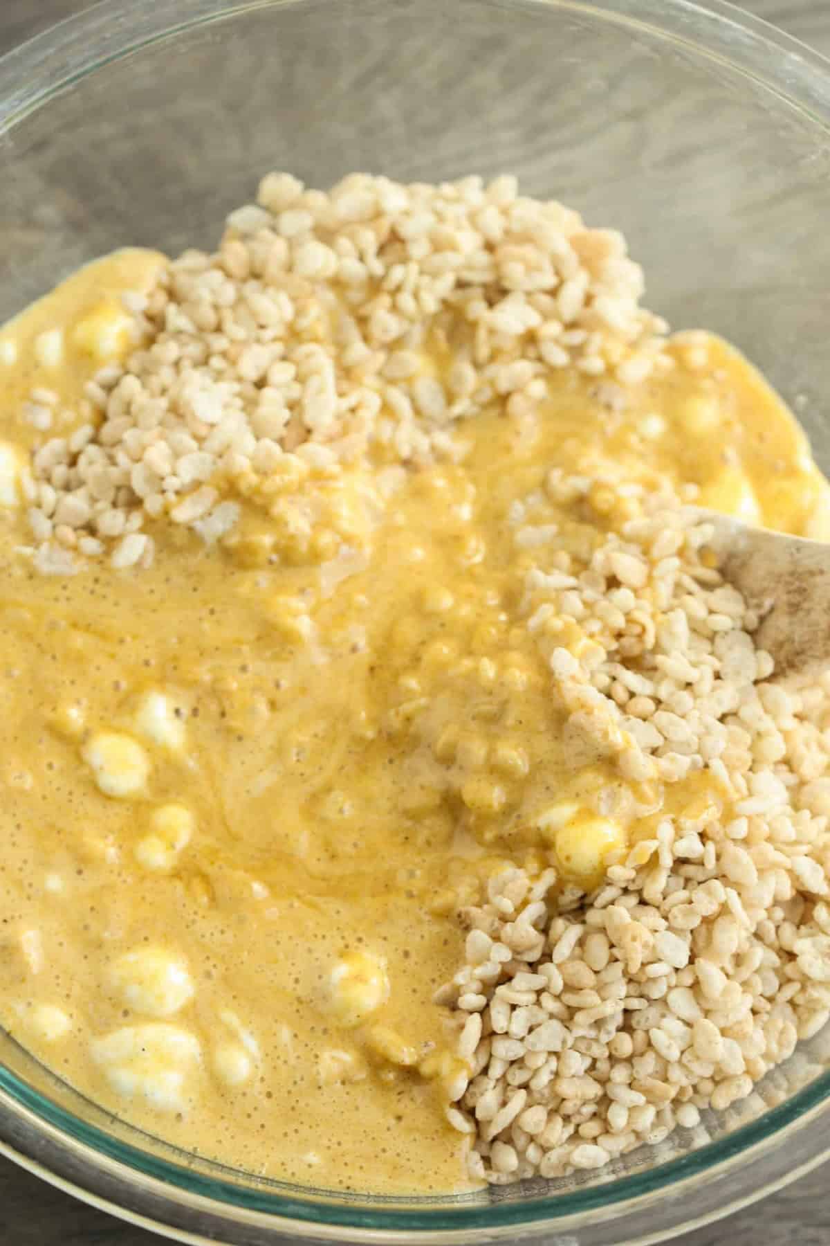 A glass bowl containing a mixture of melted marshmallow, cereal, and possibly pumpkin puree being stirred with a wooden spoon.