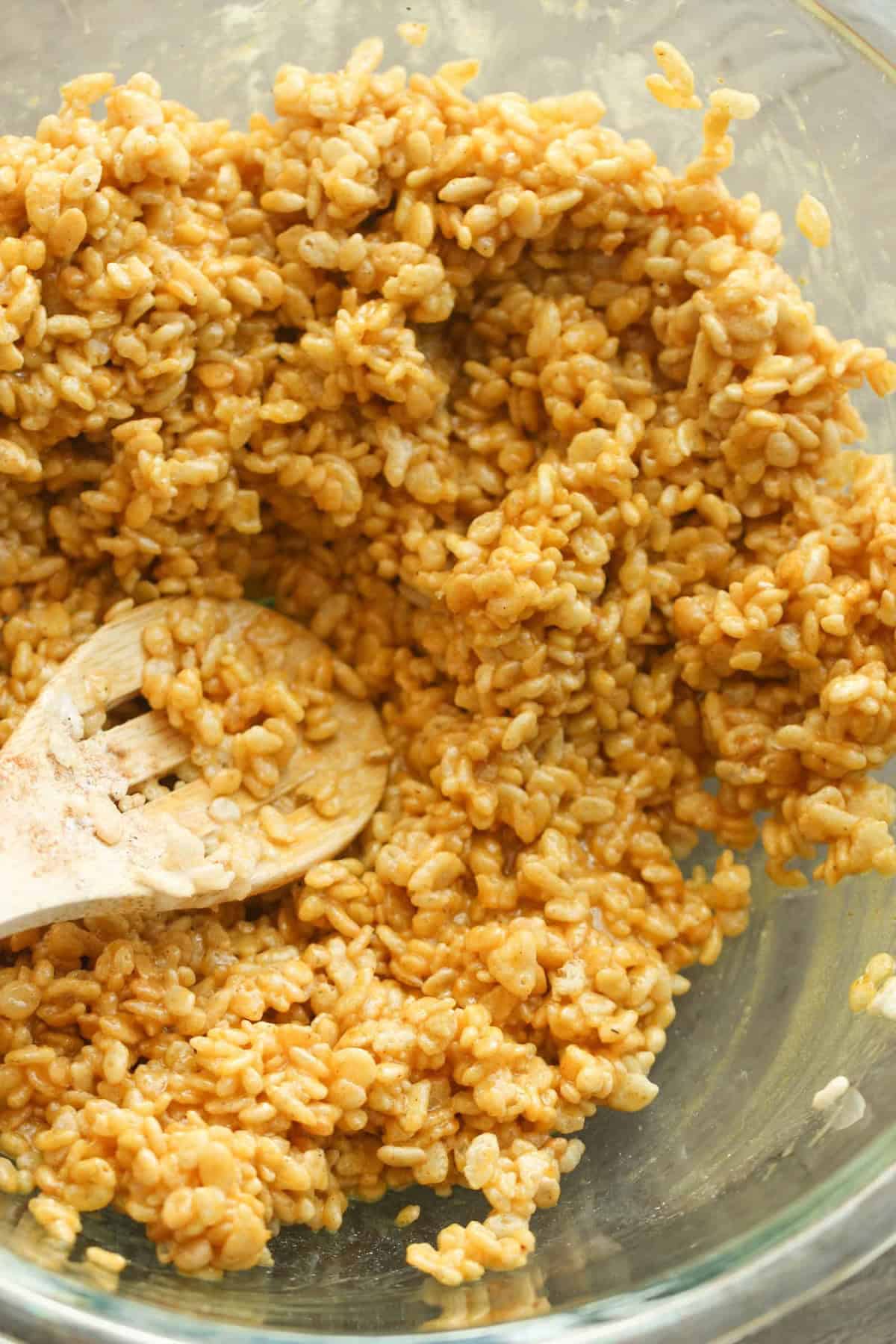 A wooden spoon stirs a mixture of crispy rice cereal and melted marshmallows in a glass bowl.