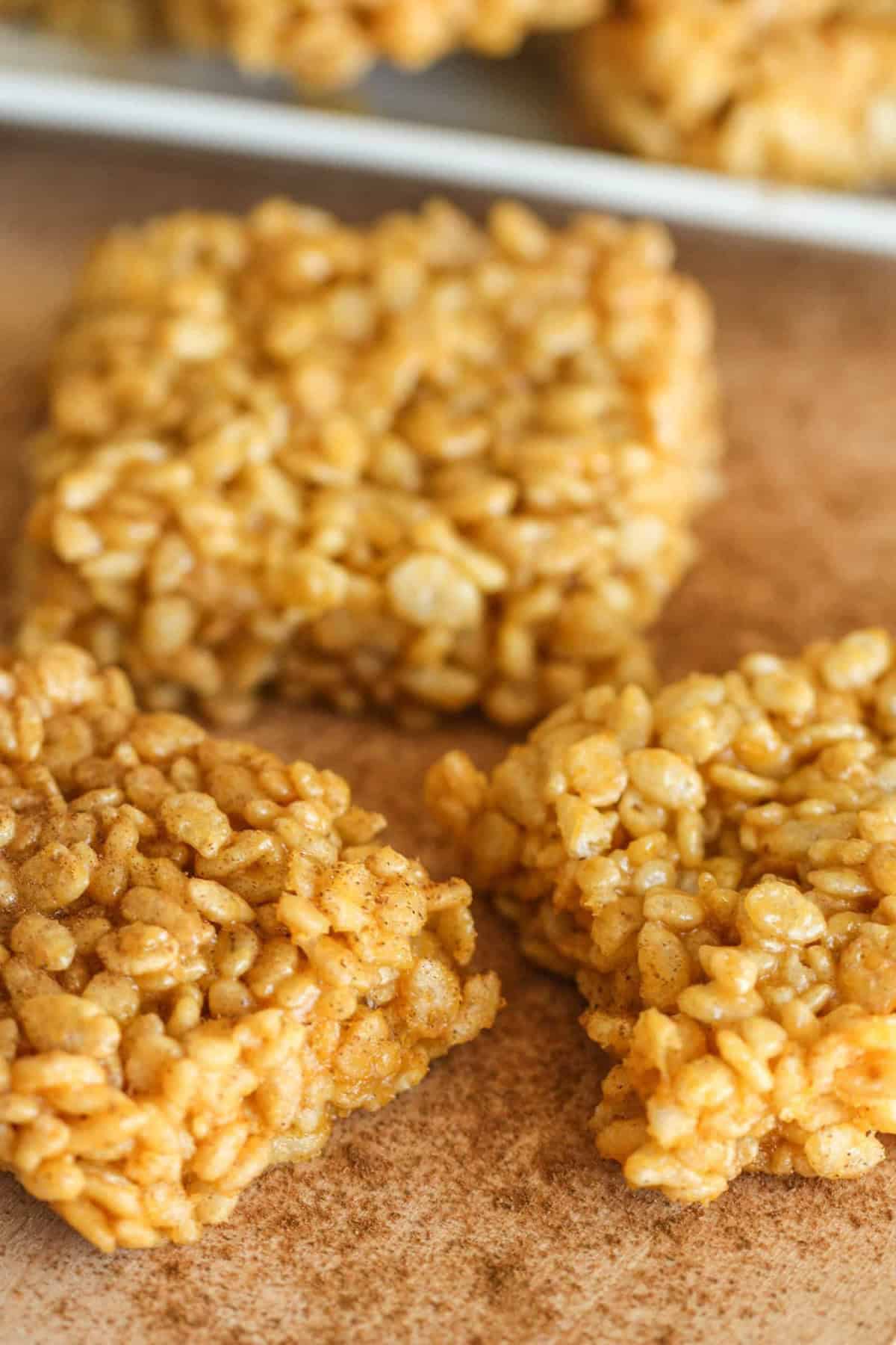 Close-up of three square rice crispy treats on a brown surface. The treats have a golden color and a crunchy texture.