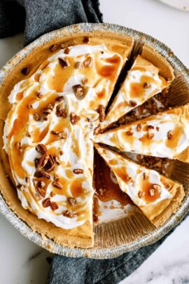 A pie with a graham cracker crust, topped with whipped cream, caramel drizzle, and pecan pieces, is shown with several slices cut.