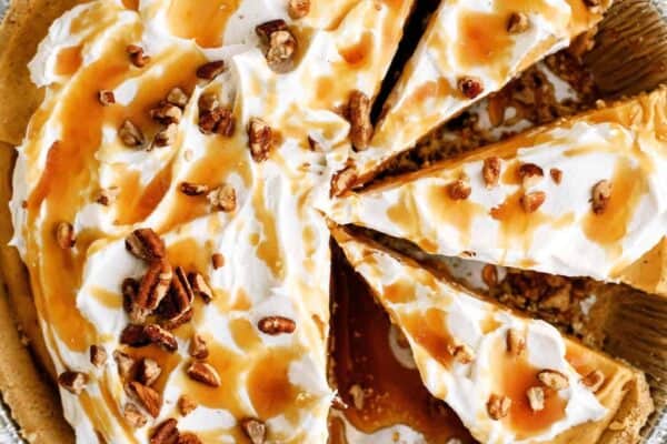 A pie with a graham cracker crust, topped with whipped cream, caramel drizzle, and pecan pieces, is shown with several slices cut.