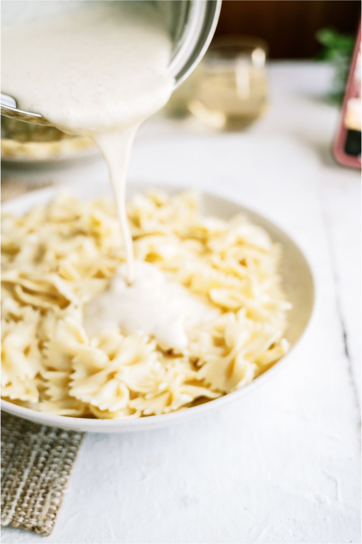 Pouring white sauce on top of a large plate filled with cooked bowtie pasta.