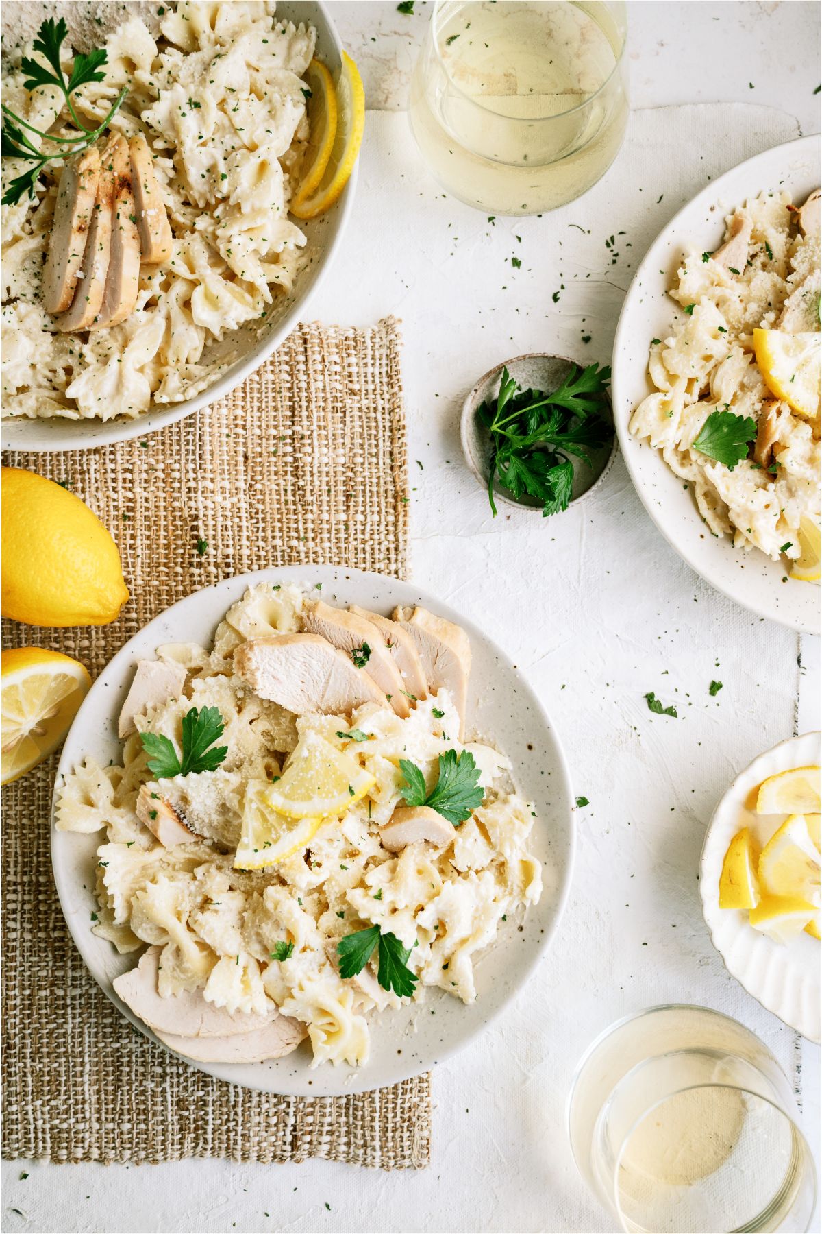 Top view of 3 plates of Lemon Pasta with sliced chicken. and sliced lemons on top. Tpw lemons on a cutting board and a small bowl of parsley on the side.