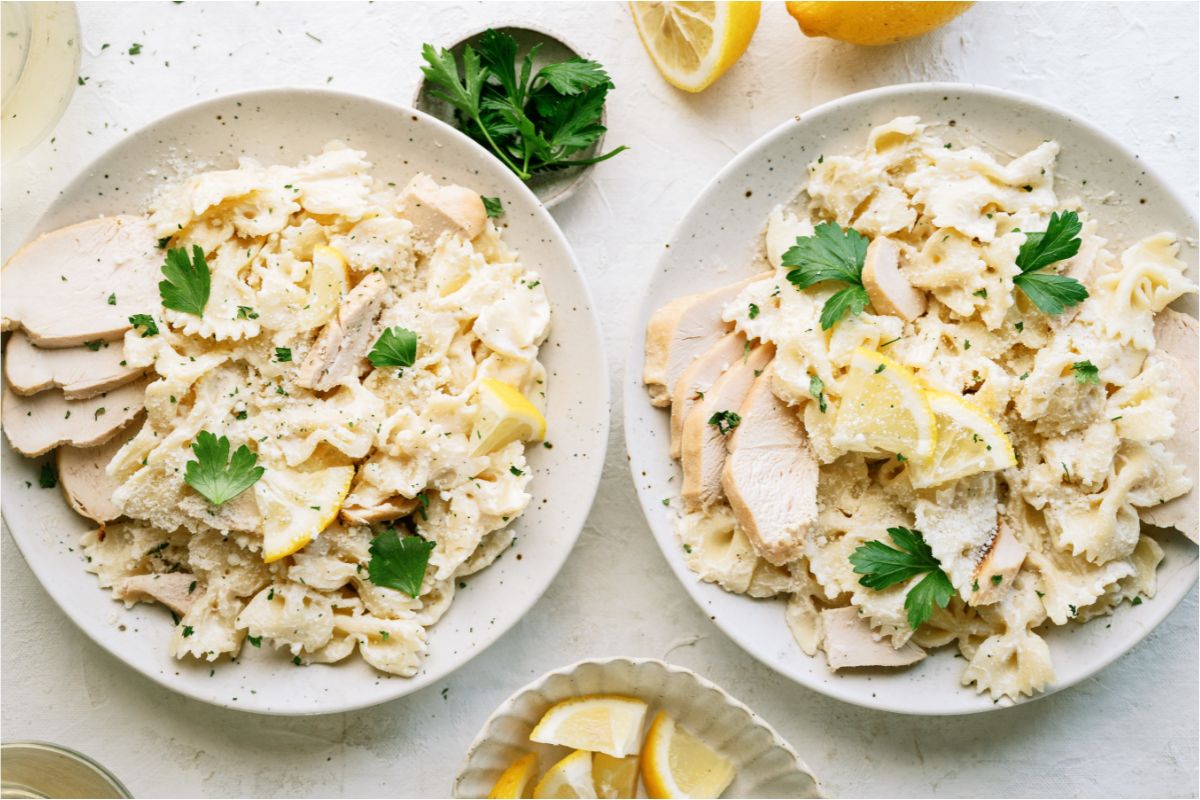 Two plates side by side with pasta, sliced chicken, white sauce, parsley and shredded cheese. Topped with sliced lemons. Lemons and Parsley in the background.