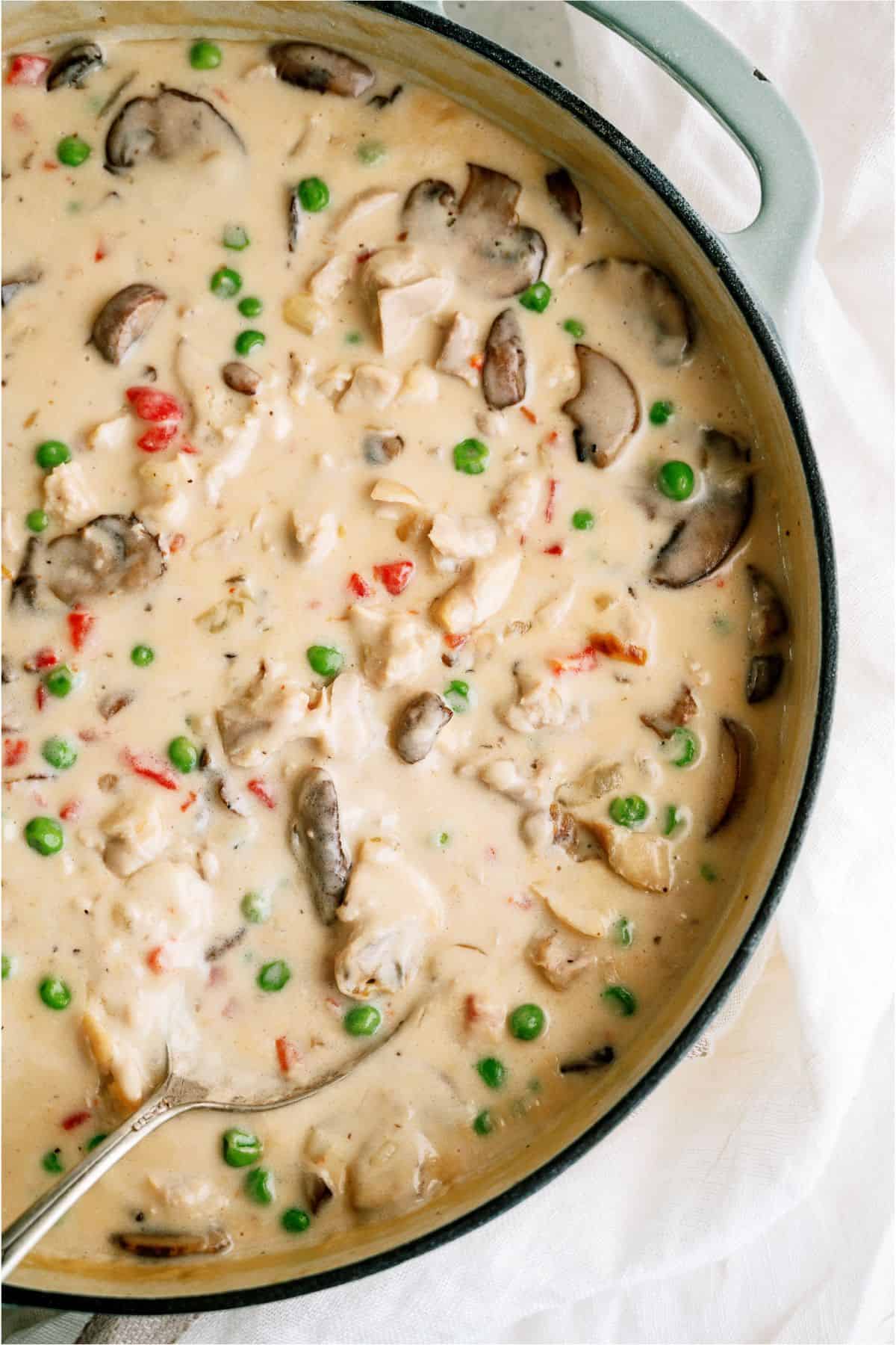 A creamy casserole dish with chicken, mushrooms, peas, and red bell peppers, topped with a sauce and a serving spoon placed inside.