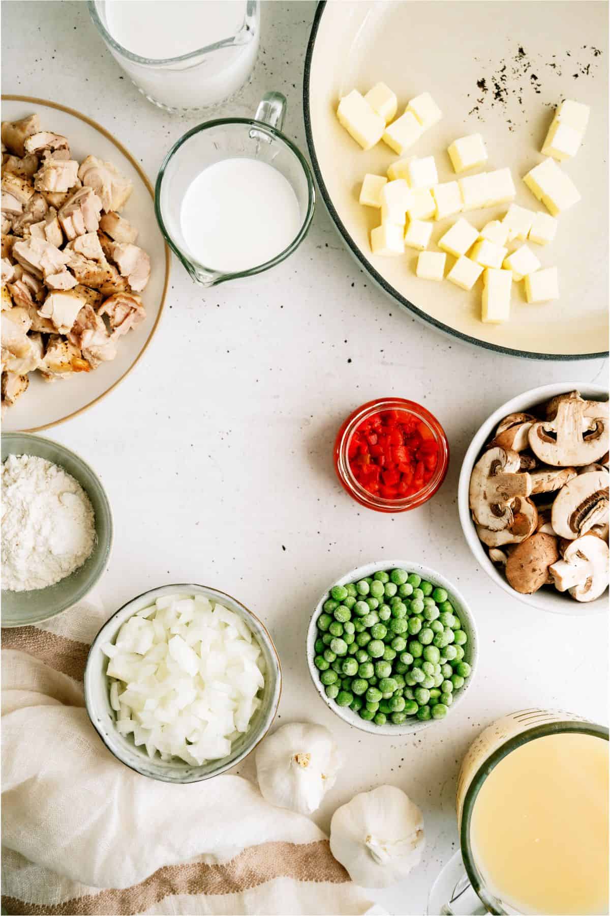 A countertop displays ingredients including diced butter, chopped onions, peas, mushrooms, diced garlic, diced chicken, flour, milk, cream, diced red bell peppers, and chicken broth.