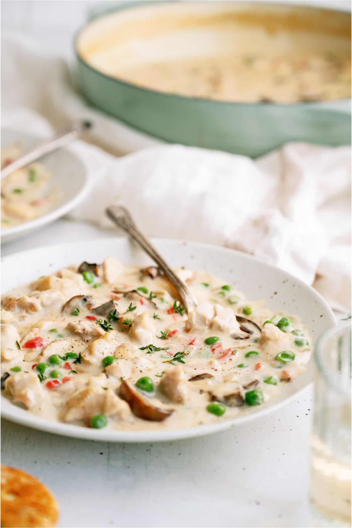 A bowl of creamy chicken and vegetable stew with peas, mushrooms, and red bell peppers. A spoon is rested inside the bowl, and a large dish of the stew is visible in the background.