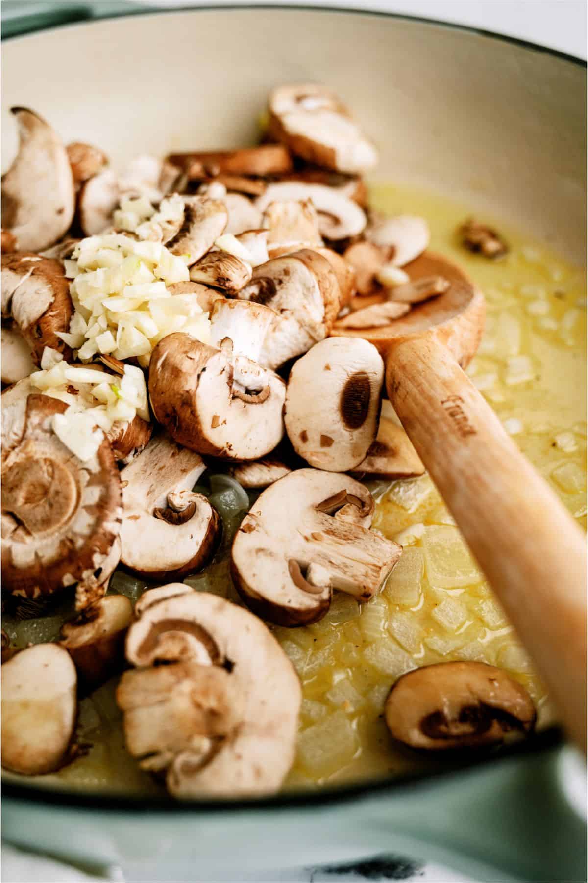 Sliced mushrooms, chopped garlic, and onions are being sautéed with oil in a frying pan with a wooden spatula.