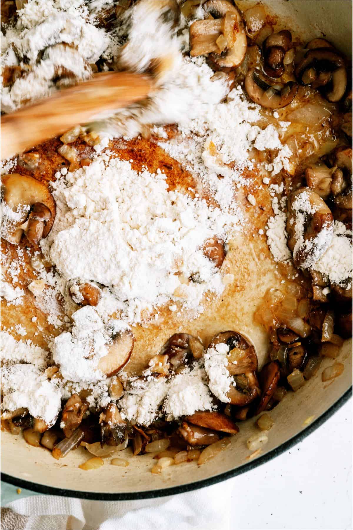 A pan of sautéed mushrooms and onions being stirred, with flour being added to the mixture.