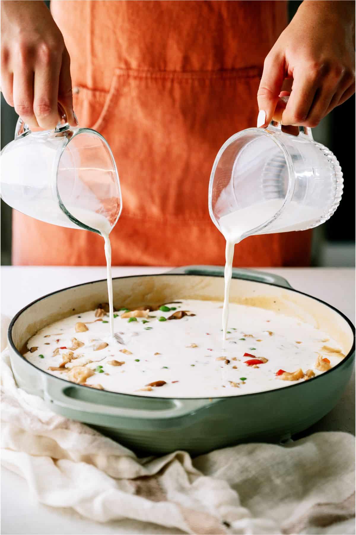 Person wearing an orange apron pouring liquid from two measuring cups into a pan with a creamy mixture containing chunks of vegetables and herbs.