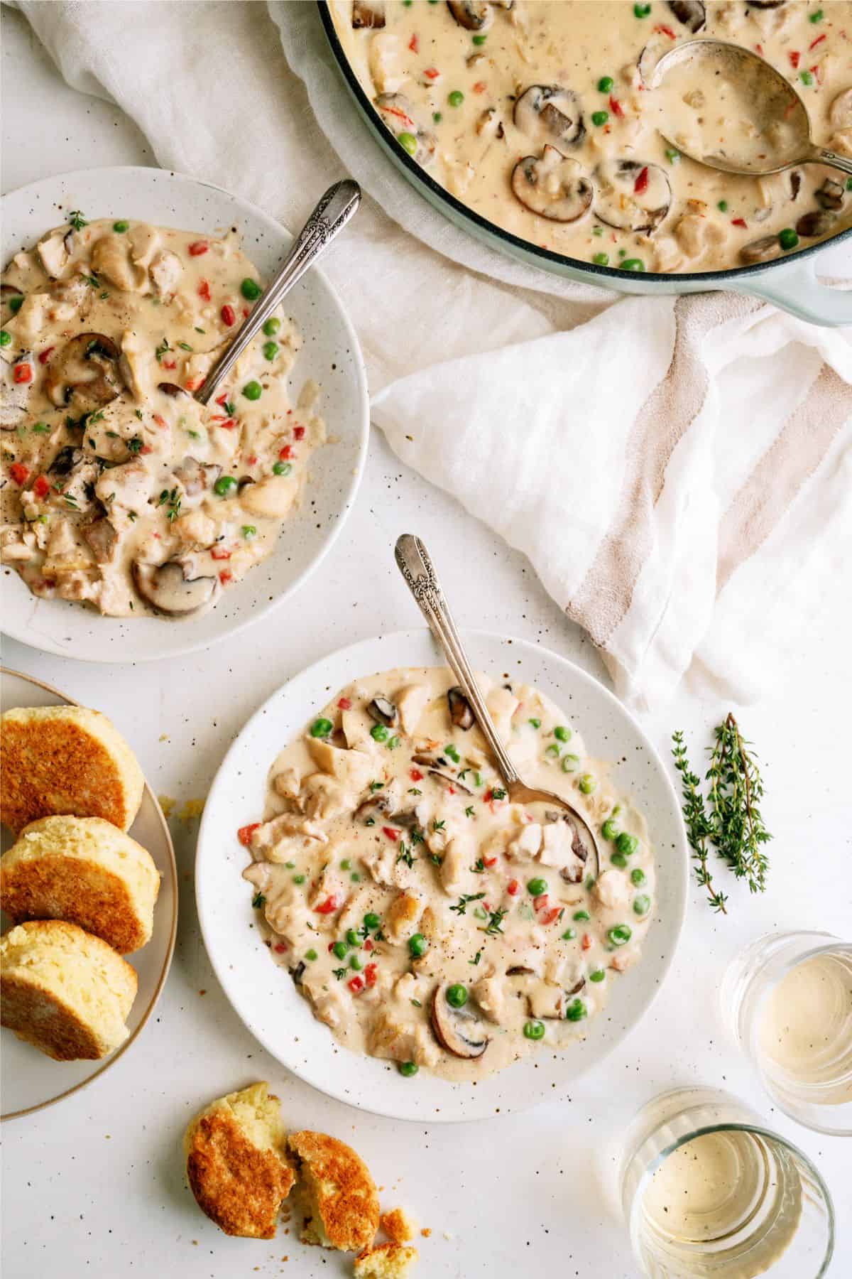 A table setting featuring bowls of creamy chicken and vegetable stew, with mushrooms, peas, and carrots. Slices of bread and two glasses of water are also on the table.