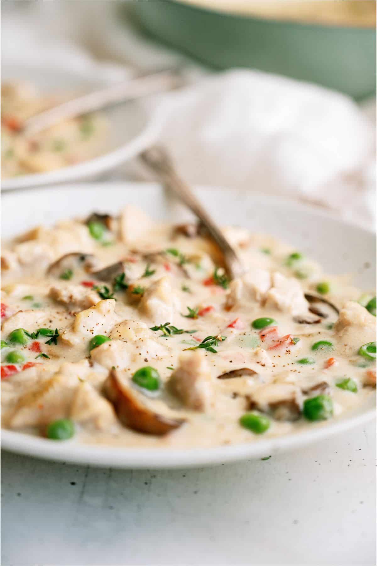 A creamy chicken and vegetable stew in a white bowl with peas, mushrooms, and red bell peppers, garnished with herbs, with a spoon in the dish.