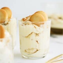 A serving of layered pudding dessert in a glass, topped with nilla wafers. Two gold spoons stacked near the bottom of the glass.