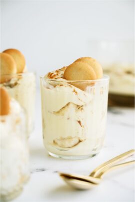 A serving of layered pudding dessert in a glass, topped with nilla wafers. Two gold spoons stacked near the bottom of the glass.