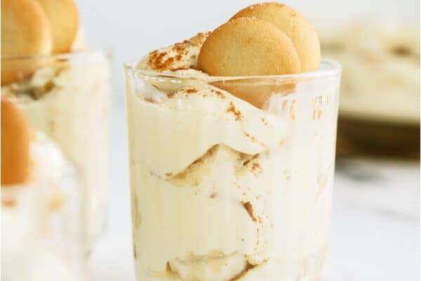 A serving of layered pudding dessert in a glass, topped with nilla wafers. Two gold spoons stacked near the bottom of the glass.