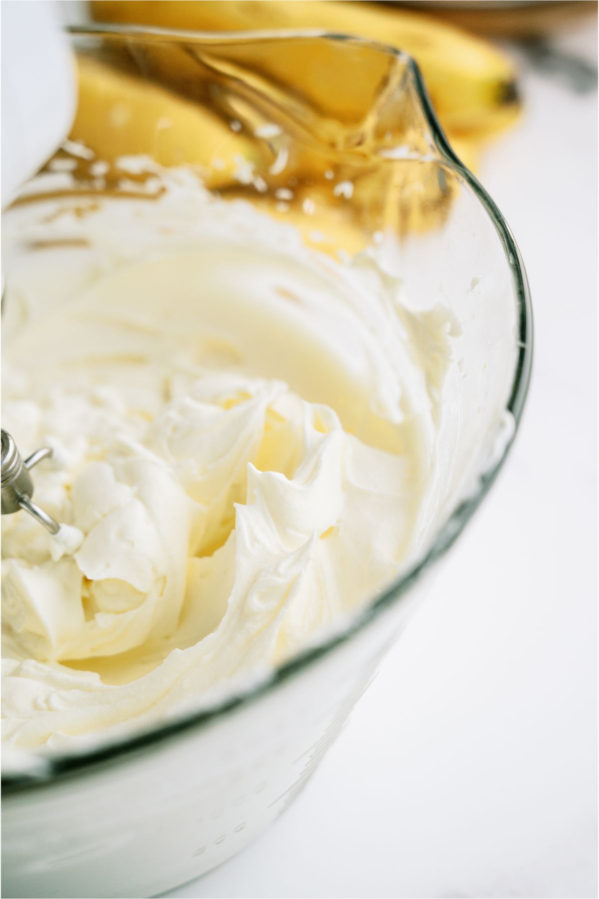 Hand mixer whipping whip cream in a glass bowl with bananas in the background.