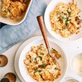 Top-down view of two bowls filled with a creamy, crumb-topped casserole dish garnished with chopped herbs, set on a light-colored table with a baking dish, wooden spoon, and glass of water nearby.