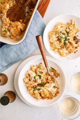 Top-down view of two bowls filled with a creamy, crumb-topped casserole dish garnished with chopped herbs, set on a light-colored table with a baking dish, wooden spoon, and glass of water nearby.