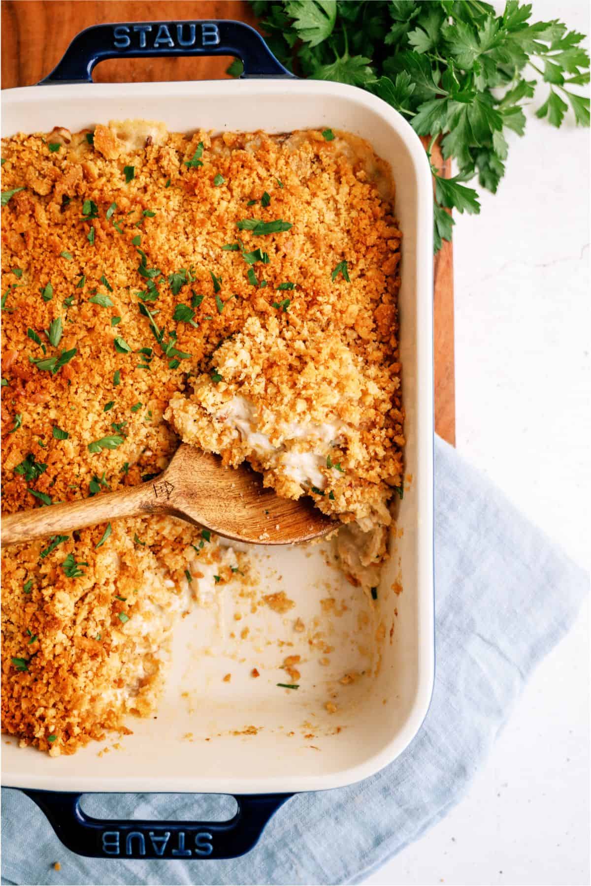 A baked dish with a breadcrumb topping is shown in a white rectangular pan. A wooden serving spoon rests inside, indicating a portion has been scooped out. Fresh parsley is in the background.