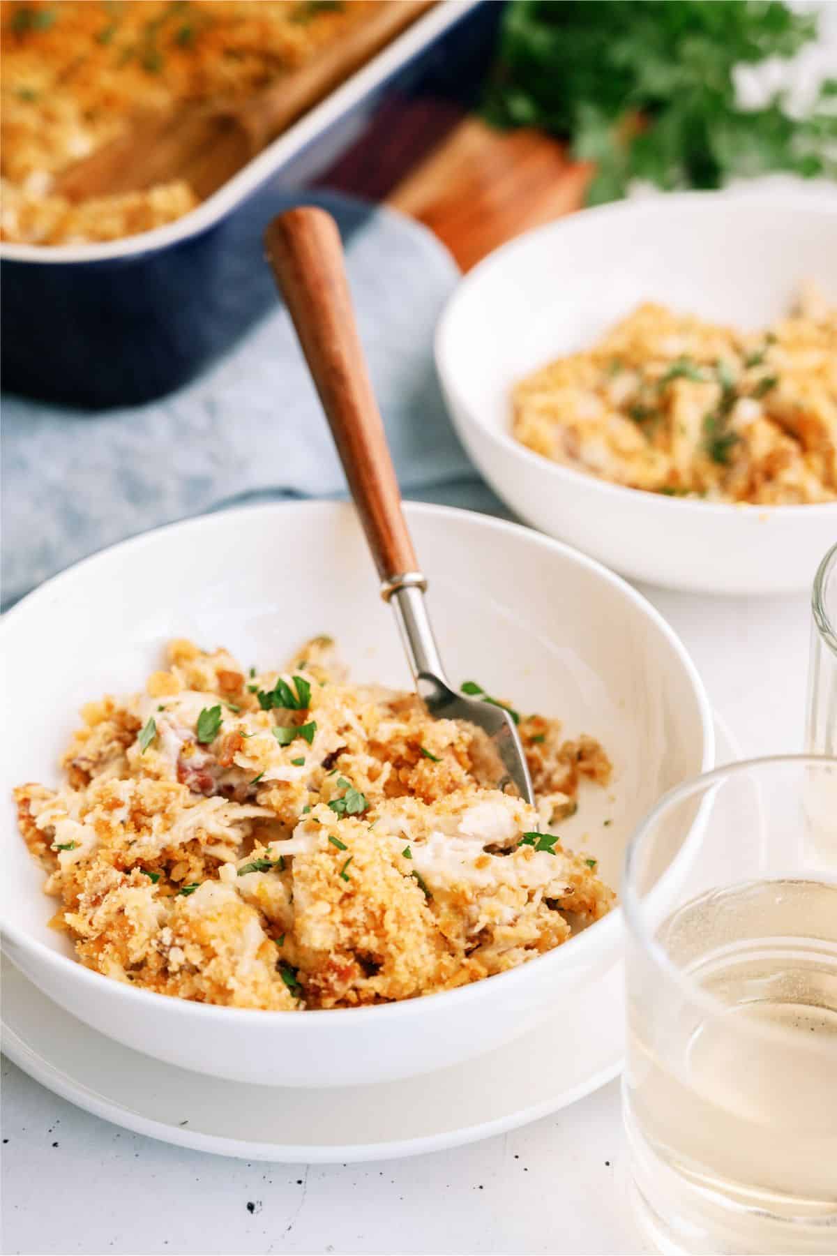 Two white bowls of casserole topped with breadcrumbs and chopped herbs, with a wooden-handled fork in one bowl. A glass of white liquid is next to the bowl and a casserole dish is in the background.