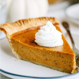 A slice of homemade pumpkin pie on a white plate with a dollop of whip cream on top.