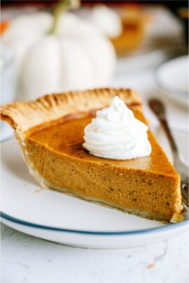 A slice of homemade pumpkin pie on a white plate with a dollop of whip cream on top.