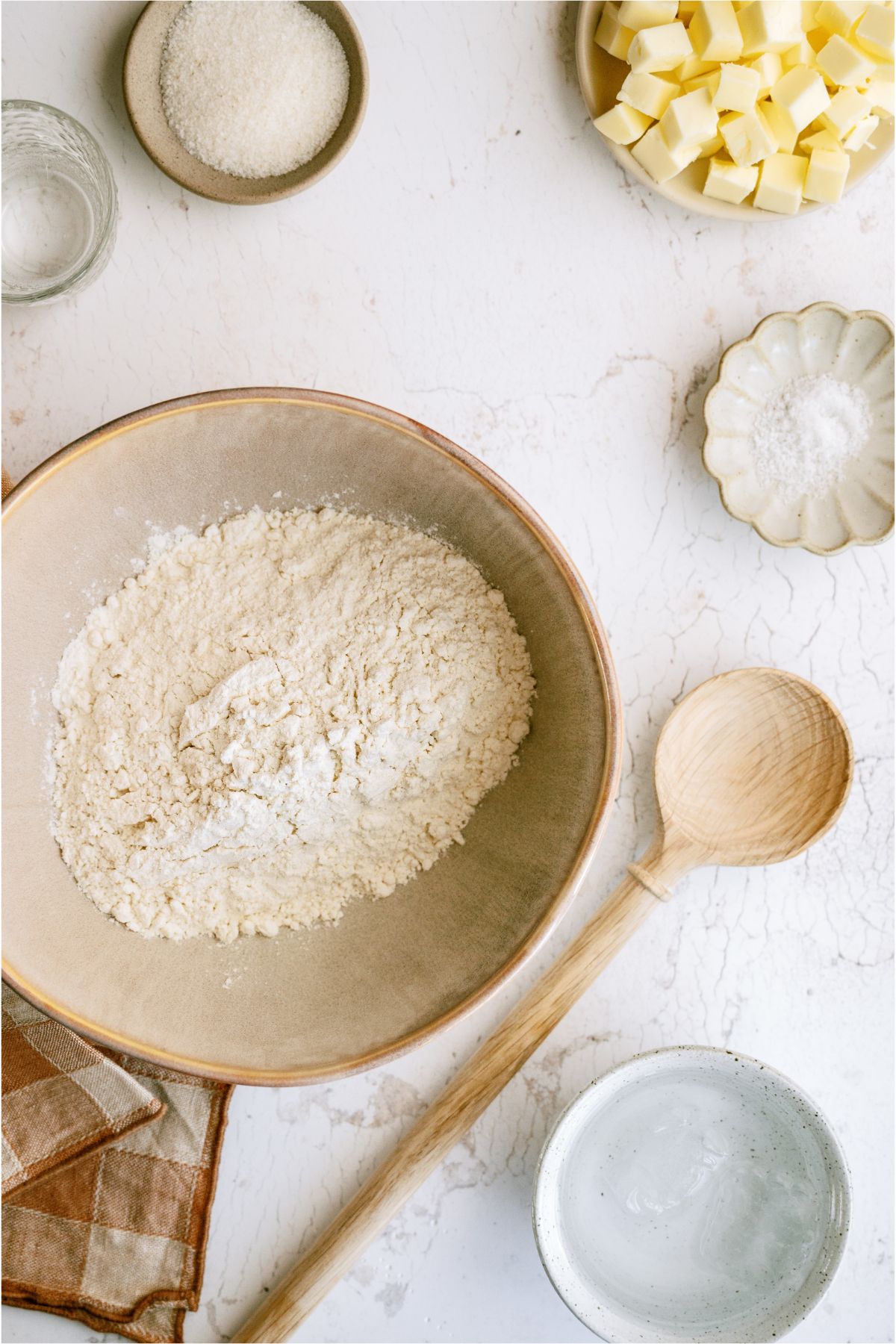 A mixing bowl with flour, small bowls with other dry ingredients, a bowl of cubed butter and a wooden spoon.