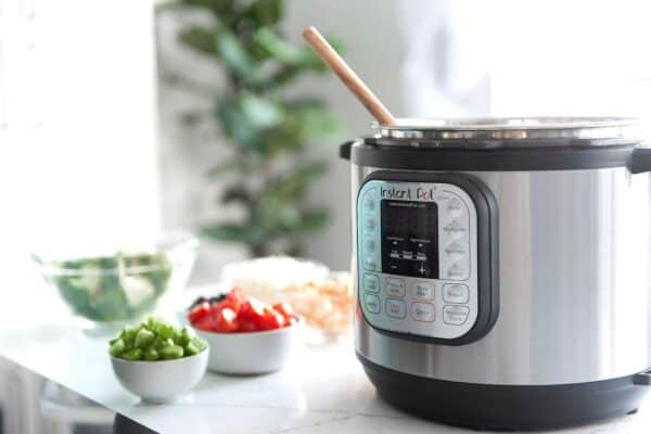 An Instant Pot on a counter with a wooden spoon leaning inside. In the background, chopped vegetables are placed in bowls next to the appliance.