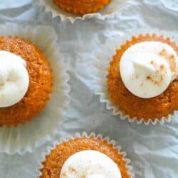 Five pumpkin cupcakes with dollops of white frosting dusted with cinnamon, placed on a crinkled white surface.