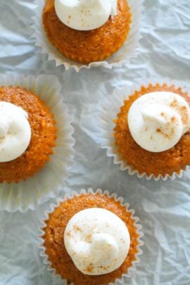 Five pumpkin cupcakes with dollops of white frosting dusted with cinnamon, placed on a crinkled white surface.