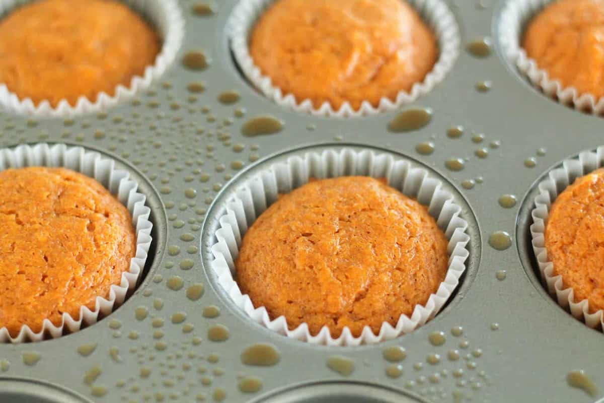 Close-up of six freshly baked muffins in a silver muffin tray, each lined with white paper cups and with droplets of moisture on the tray.