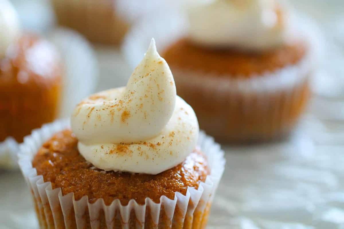 A close-up of a cupcake with cream cheese frosting and a sprinkle of cinnamon on top. Other cupcakes are blurred in the background.