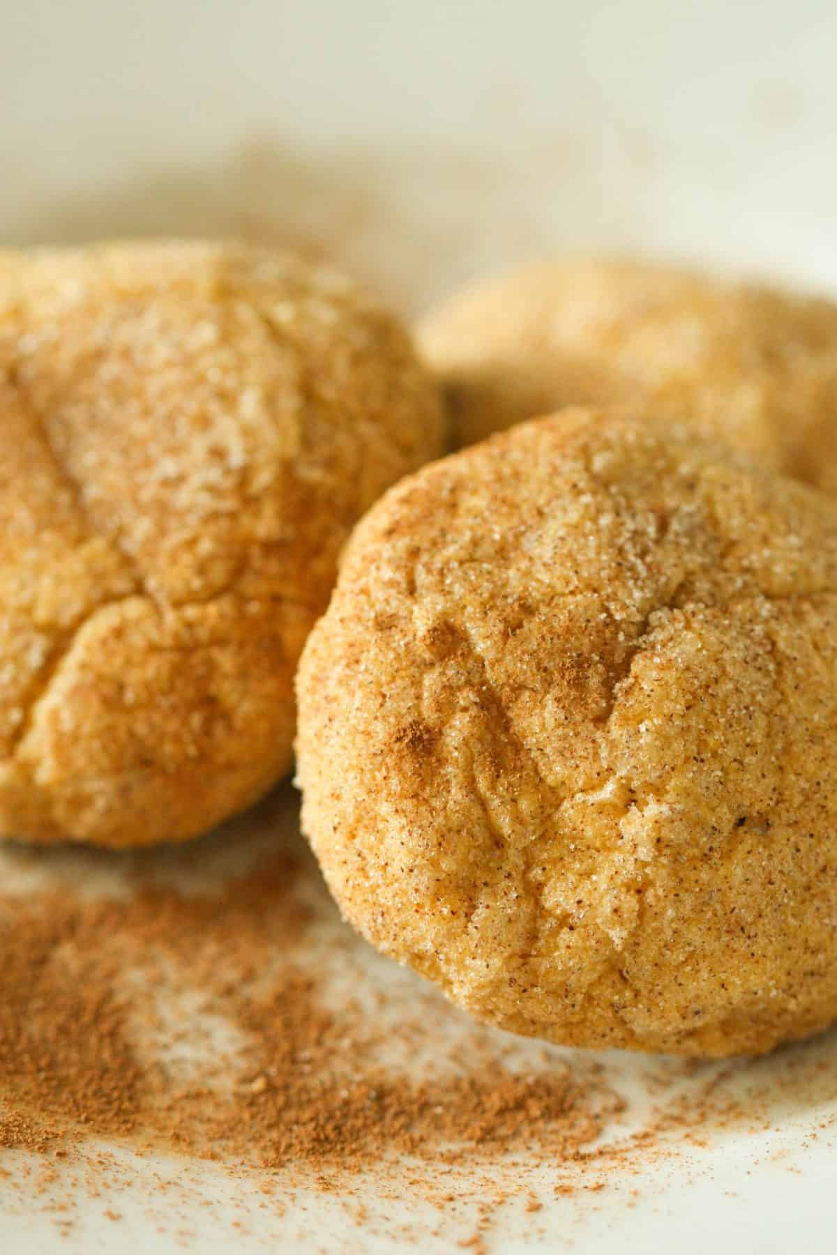 Close-up of three cinnamon-coated cookies on a white surface, with a dusting of cinnamon powder around them.