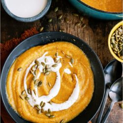 A bowl of Acorn Squash Soup with cream sauce and toasted pumpkin seeds on top. Separate bowls of Acorn Squash Soup, pumpkin seeds and cream sauce in the background.