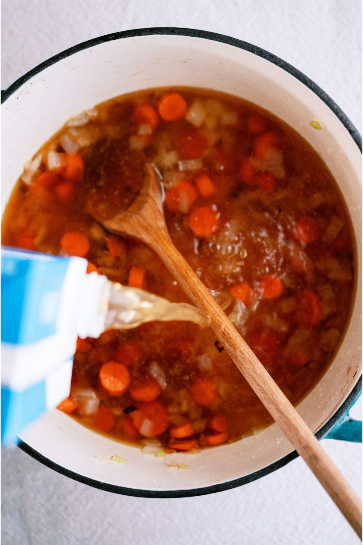 Pouring broth into a soup pot with onions and carrots and a wooden spoon.