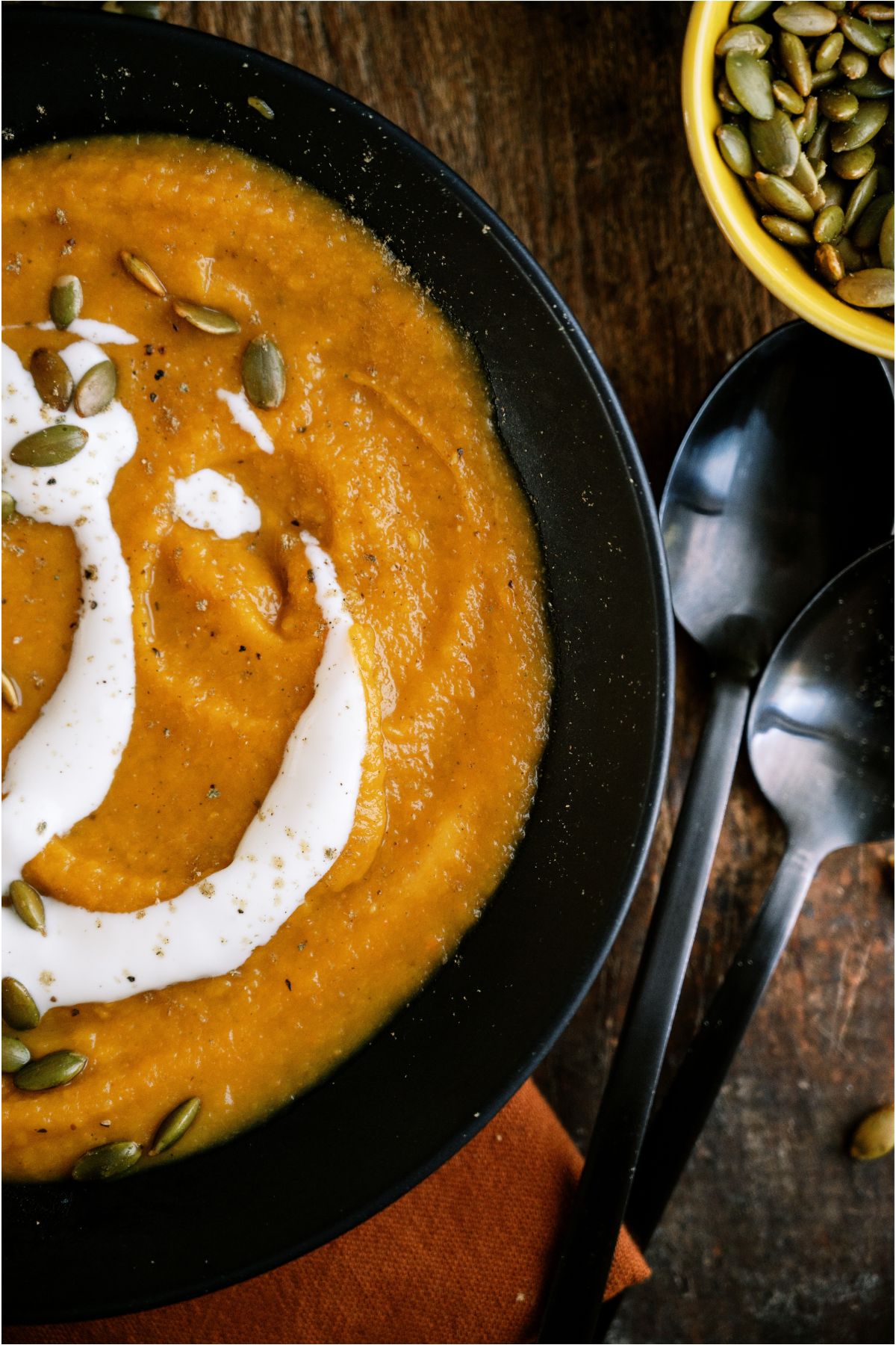 Close up of Acorn Squash Soup in a bowl topped with cream sauce and toasted pumpkin seeds with 2 spoons and a small bowl of pumpkin seeds on the side.