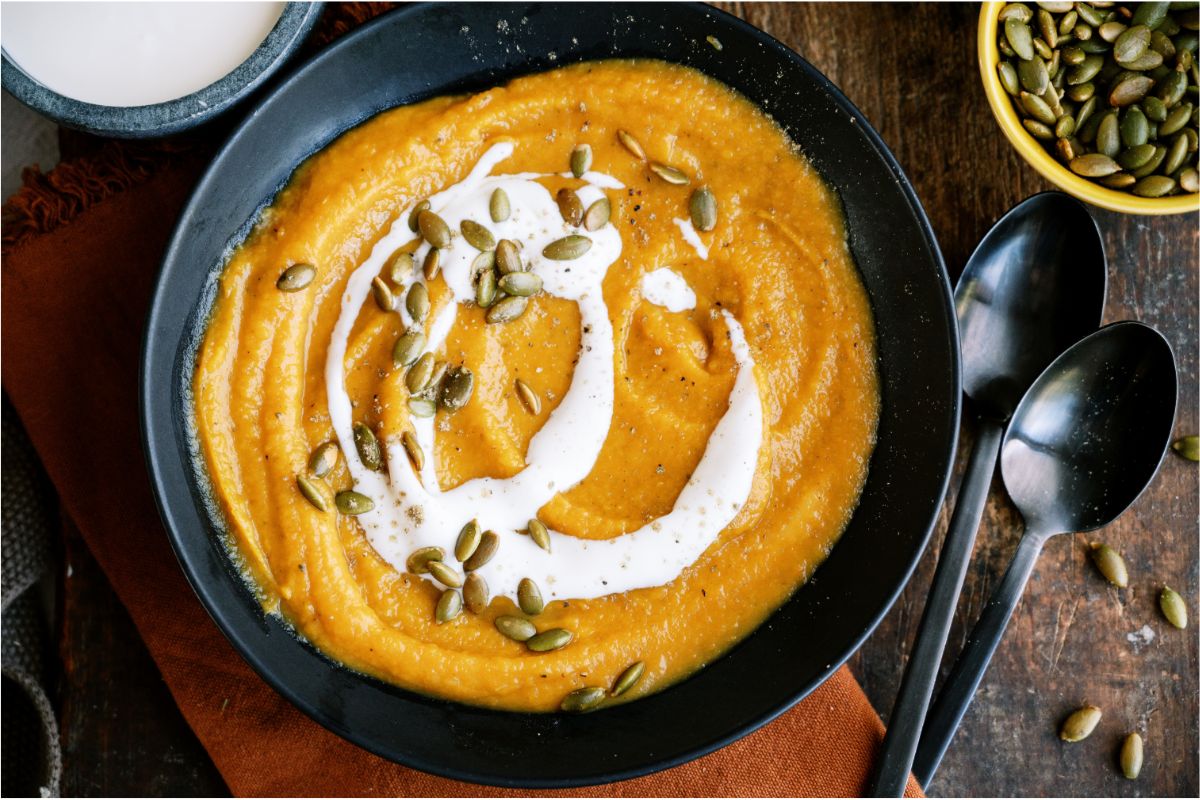 Top view of Acorn Squash Soup in a bowl topped with cream sauce and pumpkin seeds. Spoons and small bowls of cream sauce and pumpkin seeds on the side.