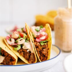 3 Cheeseburger Tacos on a plate with toppings and sauce. A jar of sauce with a spoon in it on the side.