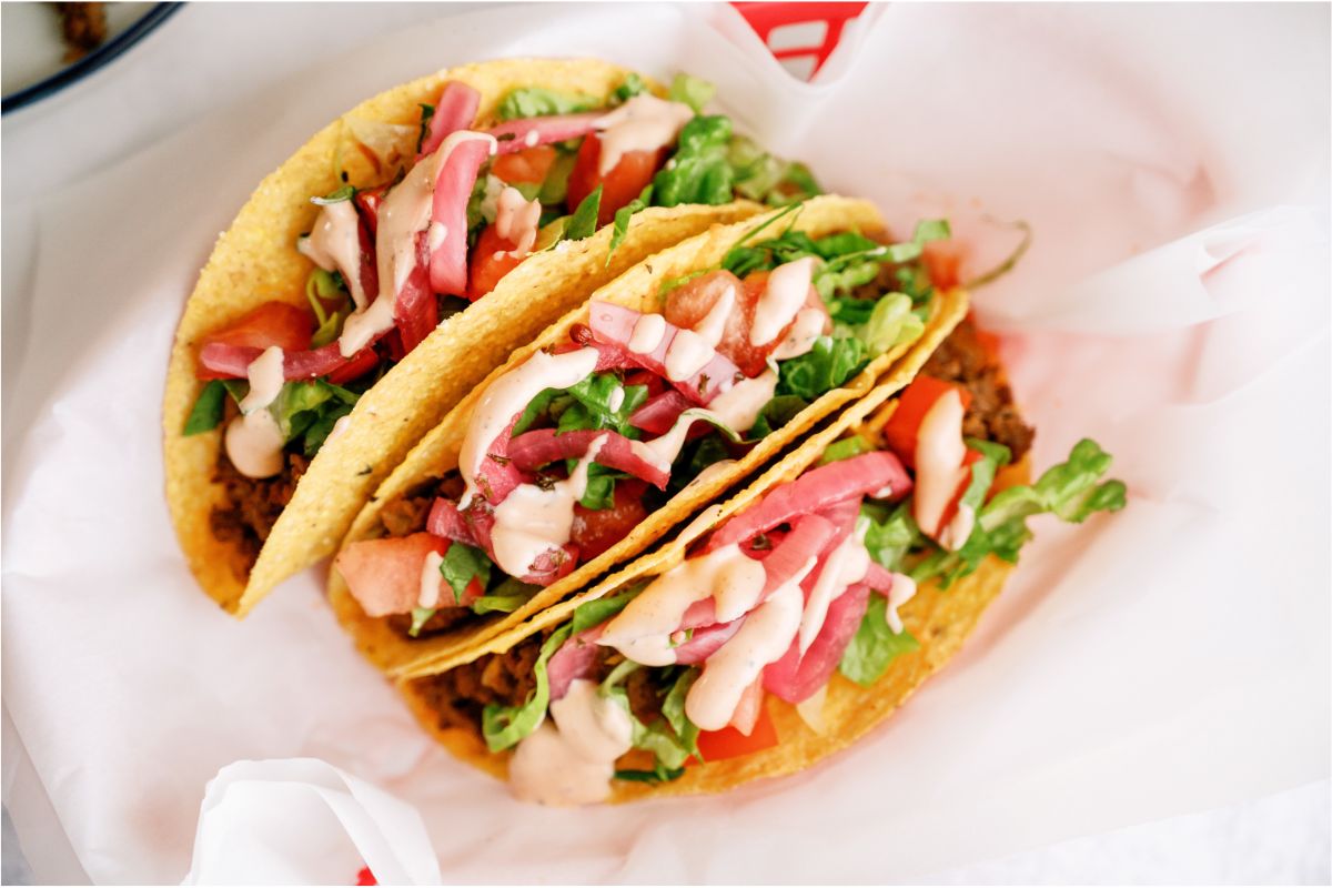 3 Cheeseburger Tacos with toppings and sauce in a red basket lined with parchment paper.