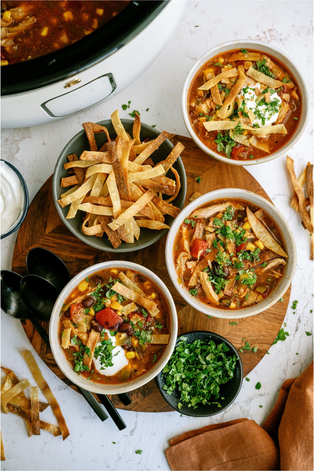 3 bowls of Easy Crock Pot Chicken Tortilla Soup on a cutting board, a bowl of tortilla strips and a bowl of chopped cilantro with the slow cooker in the background.